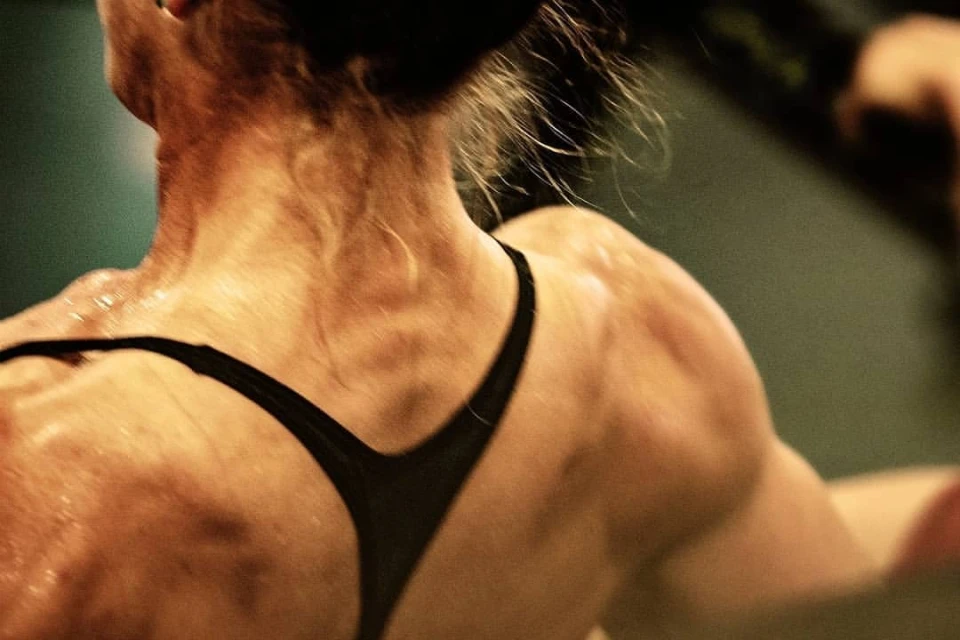 Close-up shot of the back and shoulders of an adult female athlete in a black sports bra, skin glistening with sweat in dim indoor lighting.