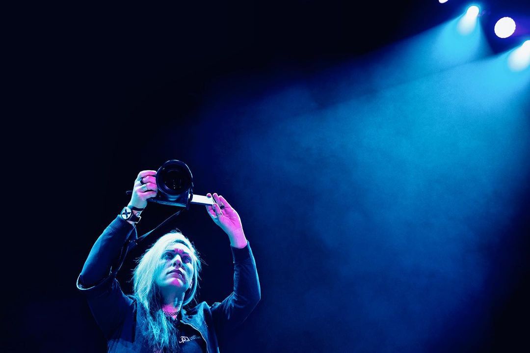 A woman in a black jacket and long blonde hair holding a DSLR camera up with blue lighting, captured in an indoor setting.