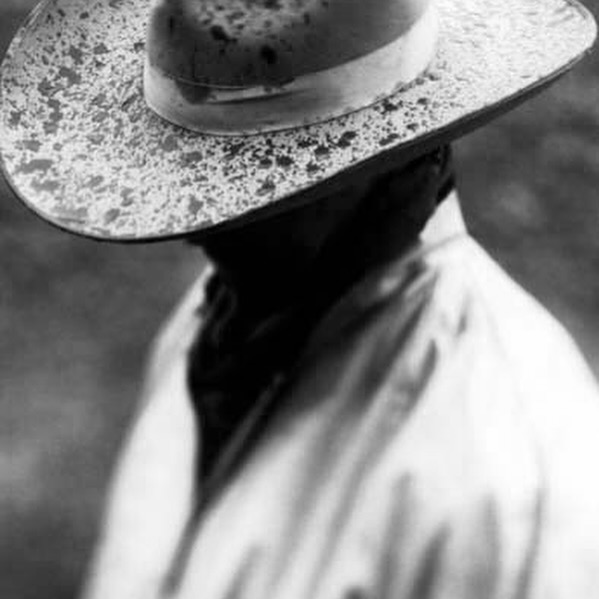Monochrome close-up shows a person in a light-colored coat and splatter-patterned hat, shoulders angled, face shadowed, and soft-focus backdrop.