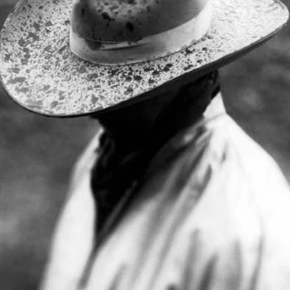 Monochrome close-up shows a person in a light-colored coat and splatter-patterned hat, shoulders angled, face shadowed, and soft-focus backdrop.