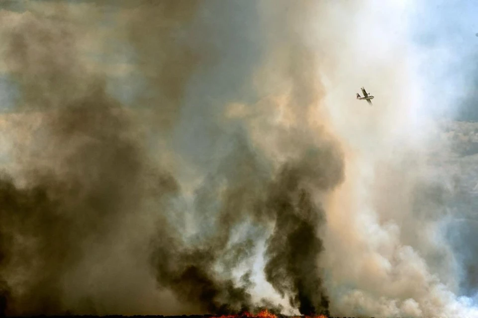 Firefighting plane flies over a wildfire, emitting smoke and flames, showcasing the aerial perspective and environmental impact.