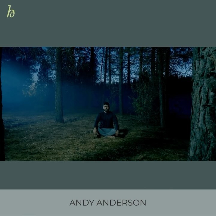 Adult man in dark sweater sits cross-legged in a forest clearing with tall trees under blue light, in a medium outdoor shot.