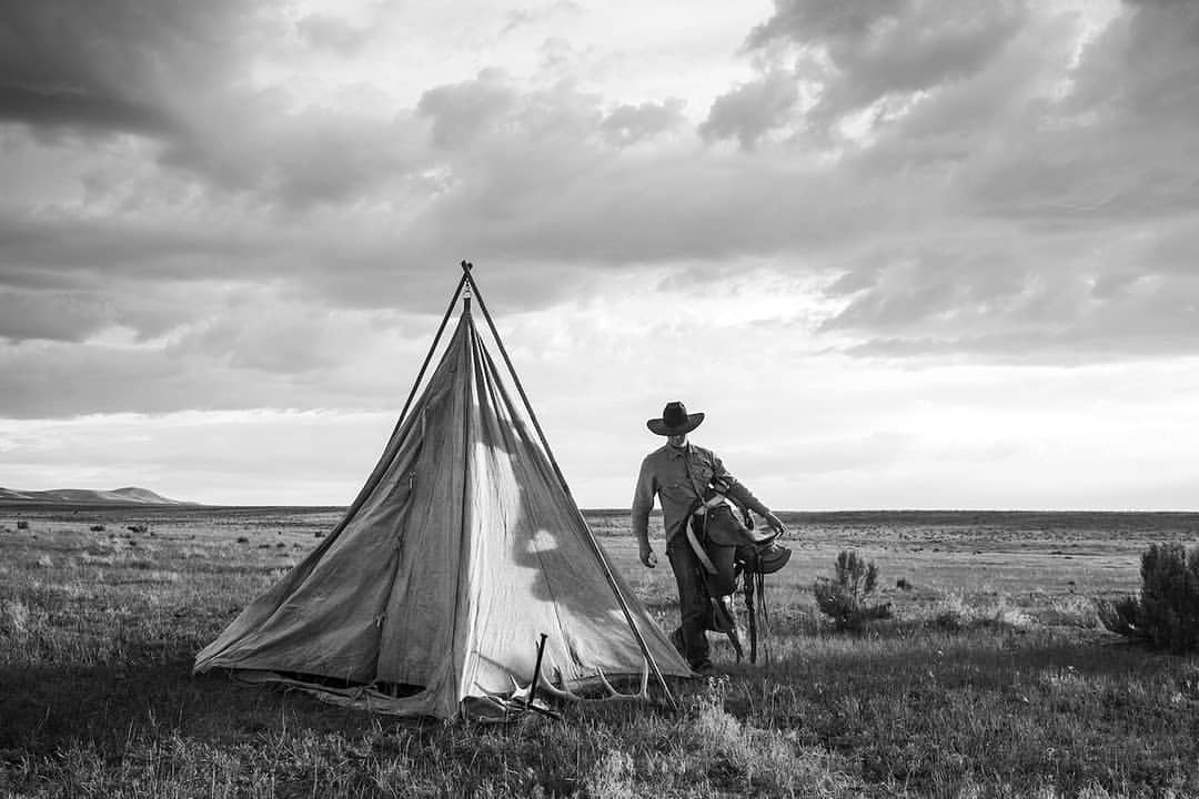 Black and white image of a cowboy standing near a tent in a rural landscape, beneath a cloudy sky, evoking a sense of the Old West.