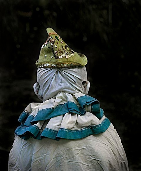 Rear view of a person wearing a light-colored ruffled collar, draped head covering, and a decorative pointed hat against a dark background.