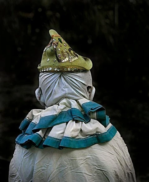 Rear view of a person wearing a light-colored ruffled collar, draped head covering, and a decorative pointed hat against a dark background.