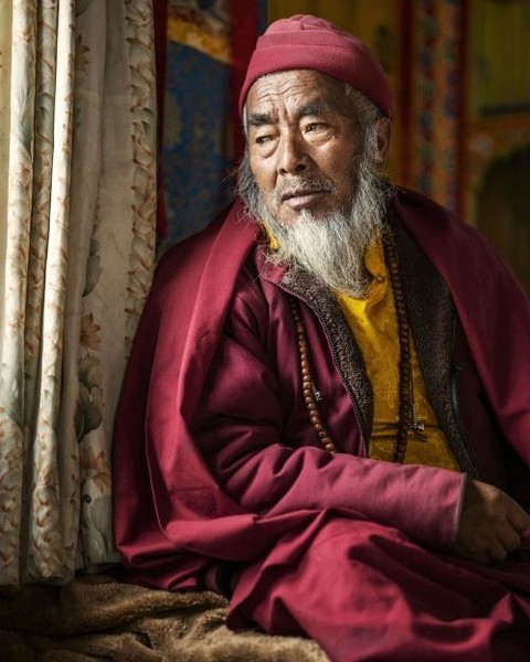 Portrait of an elderly Asian man with a white beard wearing a traditional red robe and hat, gazing contemplatively near a window.