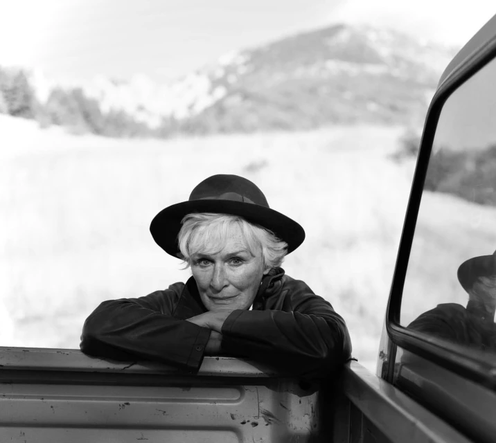 Black and white portrait of mature adult woman in dark hat and jacket, arms resting on truck bed, against a soft mountain backdrop.