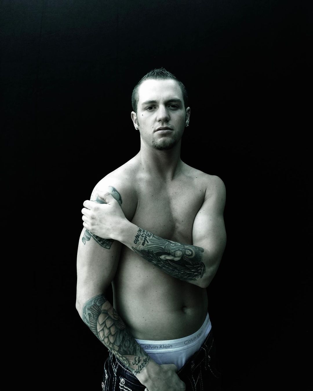 A topless Caucasian man with a mohawk haircut and arm tattoos poses in a medium studio shot against a black background.