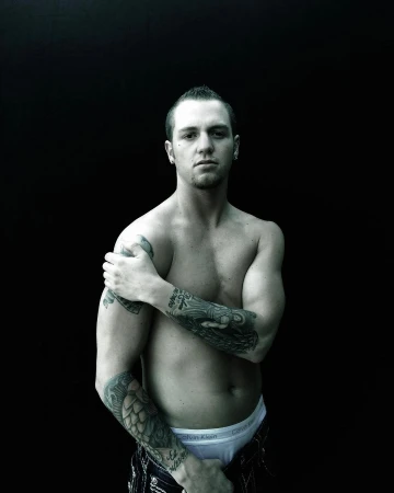 A topless Caucasian man with a mohawk haircut and arm tattoos poses in a medium studio shot against a black background.