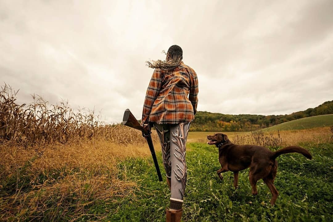 Woman in orange plaid shirt and patterned leggings walking in a field with a shotgun and a dog on a cloudy day.