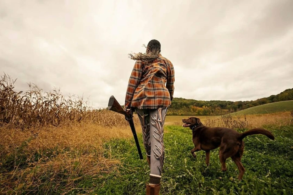 Woman in orange plaid shirt and patterned leggings walking in a field with a shotgun and a dog on a cloudy day.