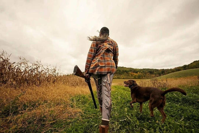 Woman in orange plaid shirt and patterned leggings walking in a field with a shotgun and a dog on a cloudy day.