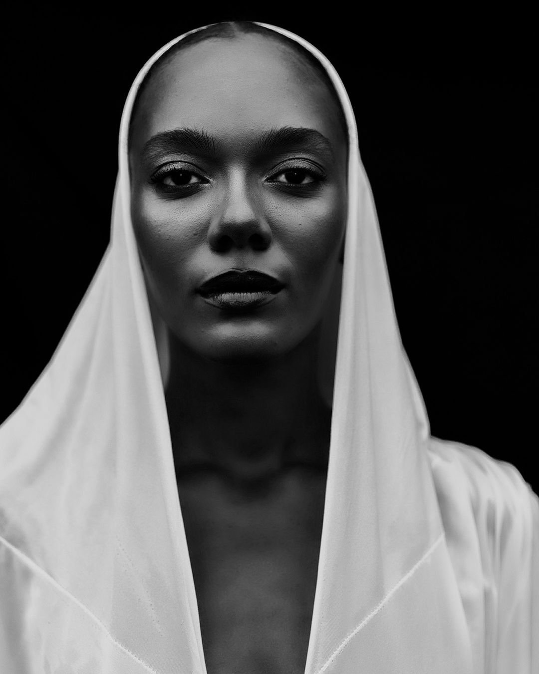 Monochrome portrait of a Black woman wearing a white hooded garment. She is gazing directly at the camera against a dark background.