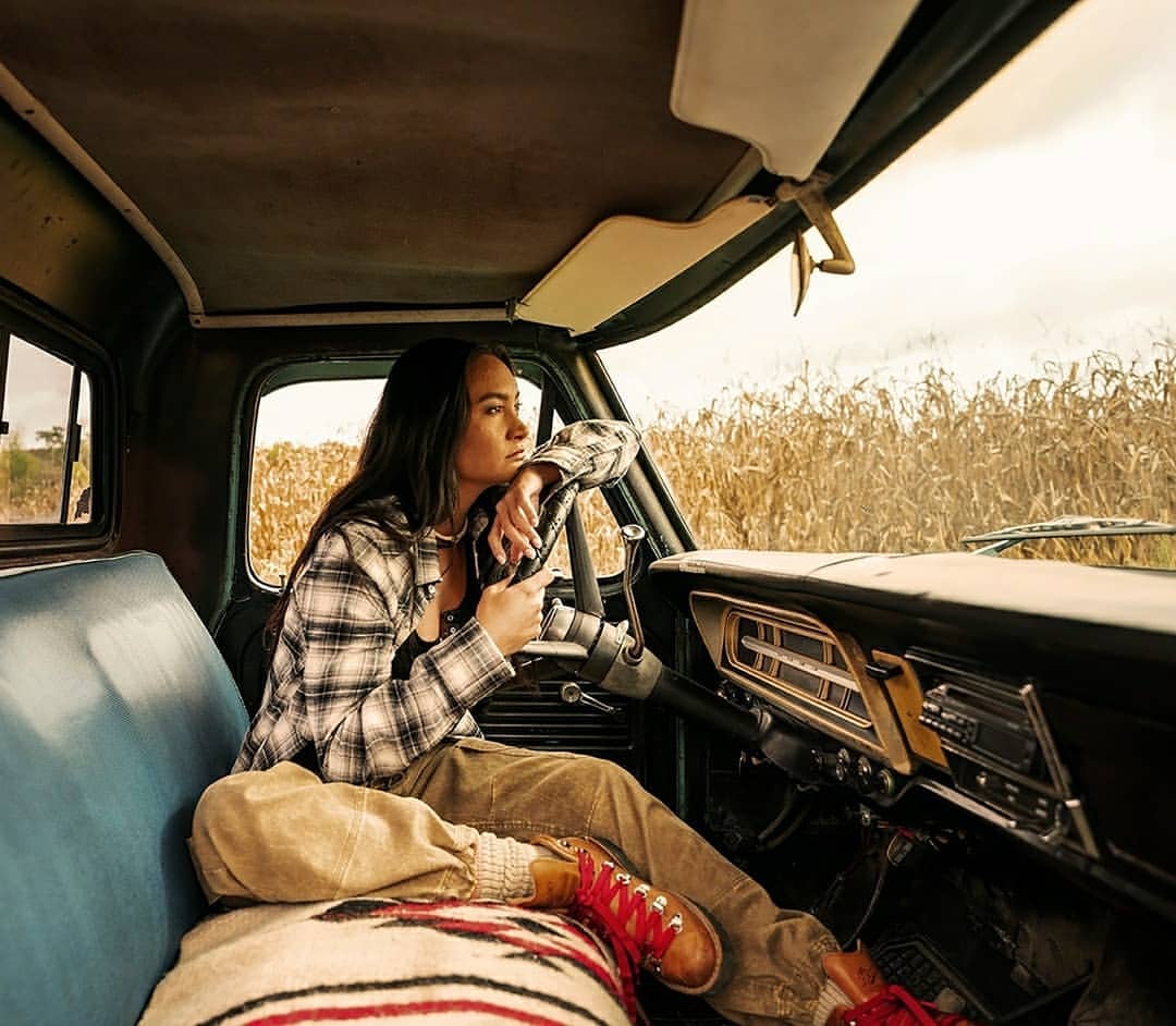 Young woman sits in a vintage truck with a blanket-covered seat and wears a plaid shirt, gazing out at a field of tall, dried corn.
