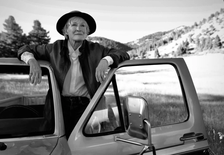 Mature woman in hat and jacket leans on pickup truck door against a mountain background in a black and white, eye-level portrait.