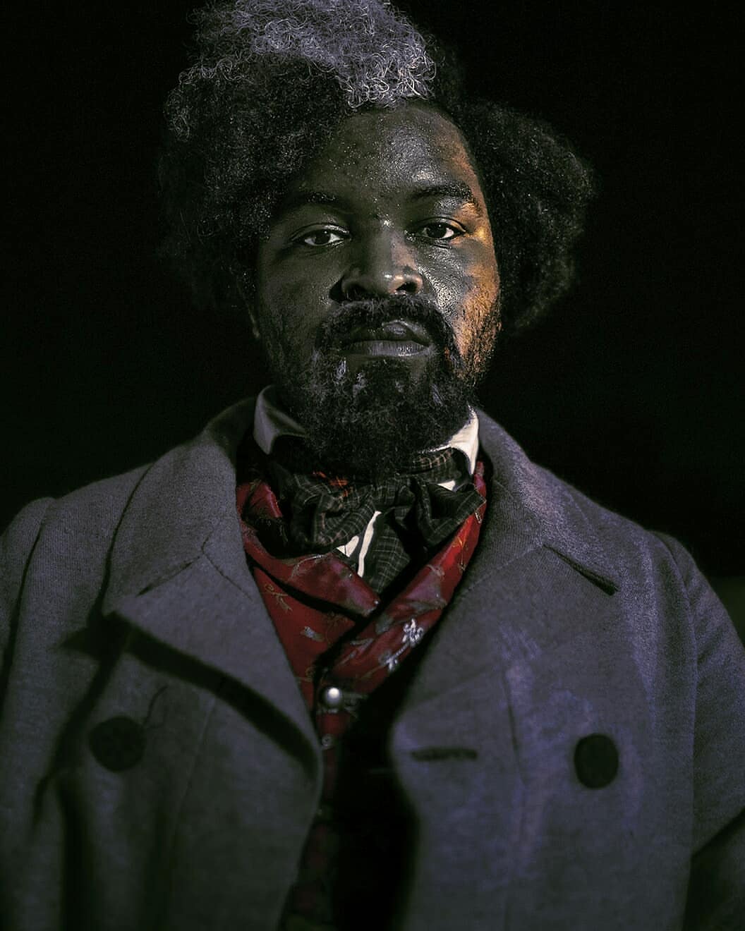 Portrait of a Black man with graying afro, wearing a Victorian-era coat and scarf, set against a dark backdrop in studio lighting.