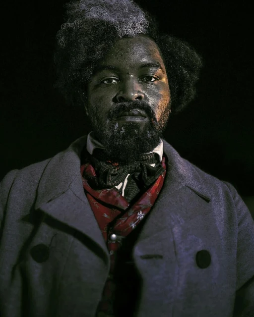 Portrait of a Black man with graying afro, wearing a Victorian-era coat and scarf, set against a dark backdrop in studio lighting.