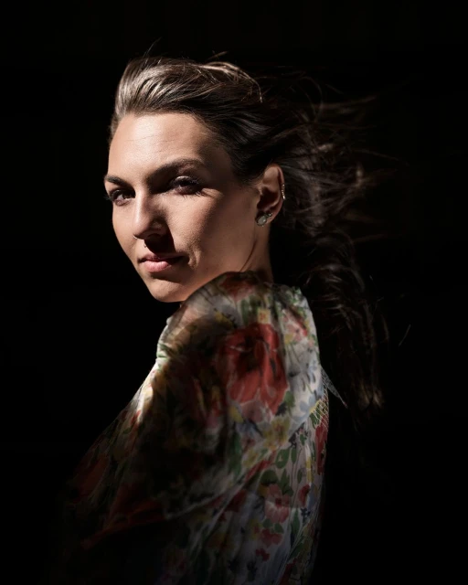 Portrait of a woman with shoulder-length brunette hair, wearing a floral blouse, posing against a dark background in studio lighting.