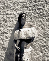 Portrait of a woman wearing an off-the-shoulder white top and dark sash standing against a textured stone wall