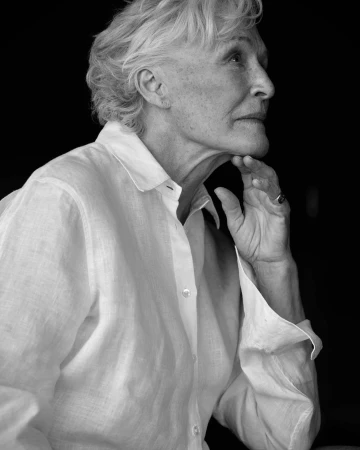 Portrait of an elderly woman with short blonde hair wearing a white button-down shirt, resting her chin on her hand against a dark background