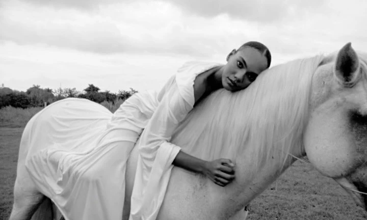 Black and white portrait of a woman in a white dress resting her head on a white horse in a field.