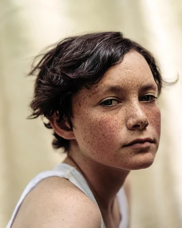 Close-up portrait of a young person with short dark hair and extensive freckles wearing a white tank top