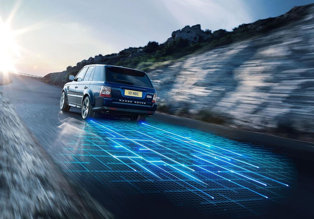 Dark blue Range Rover SUV driving on a grey winding mountain road with a geometric blue pattern below car.
