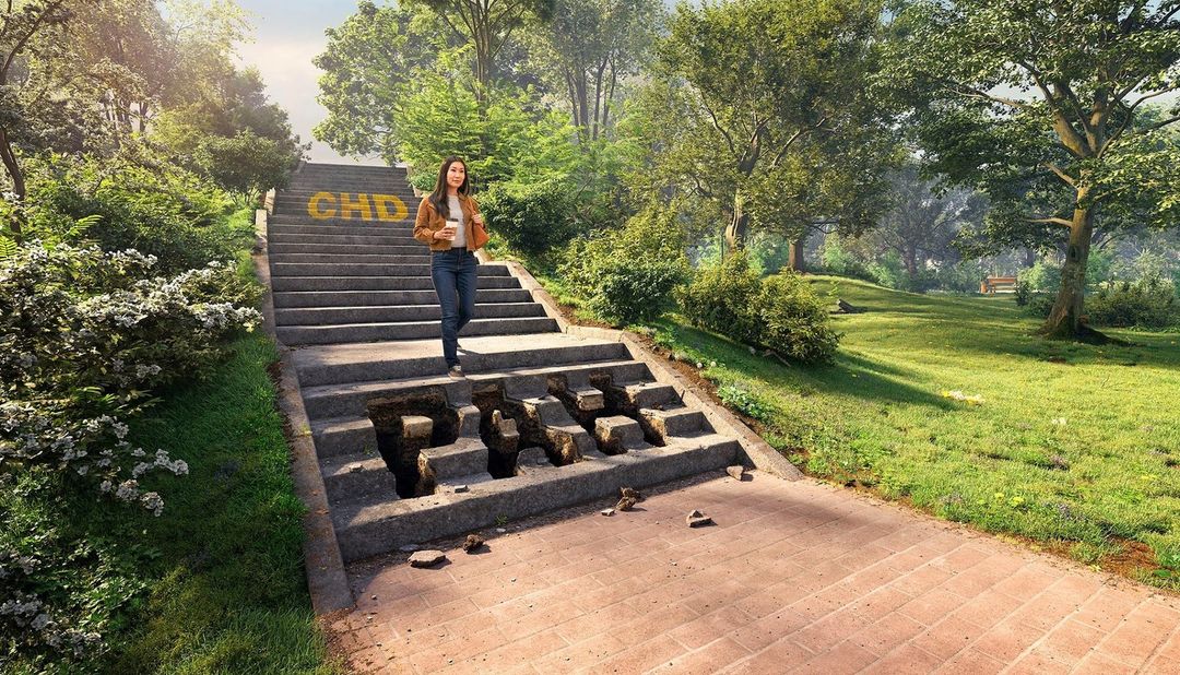 Woman walking down stone steps with missing blocks forming 'PAIN' in an outdoor park setting with green trees and grass on a sunny day.