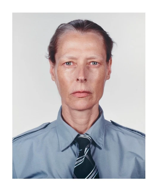 Portrait of an adult female with short grey hair wearing a blue collared shirt and tie, captured in a photo studio setting with soft lighting.