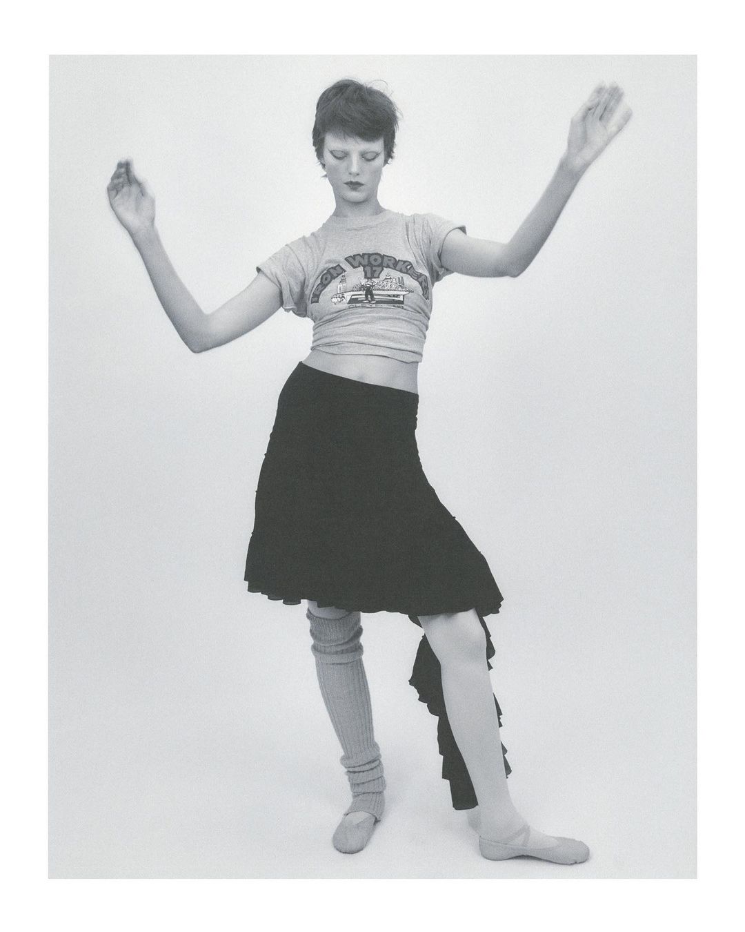 Woman in motion, wearing a t-shirt, skirt, leg warmers, and ballet slippers against a white backdrop, striking a dynamic pose with arms raised.