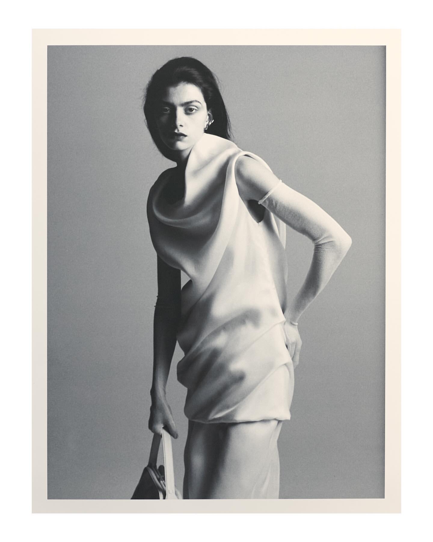 Black and white editorial portrait of a woman in a draped white dress holding a handbag against a plain grey background