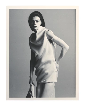 Black and white editorial portrait of a woman in a draped white dress holding a handbag against a plain grey background