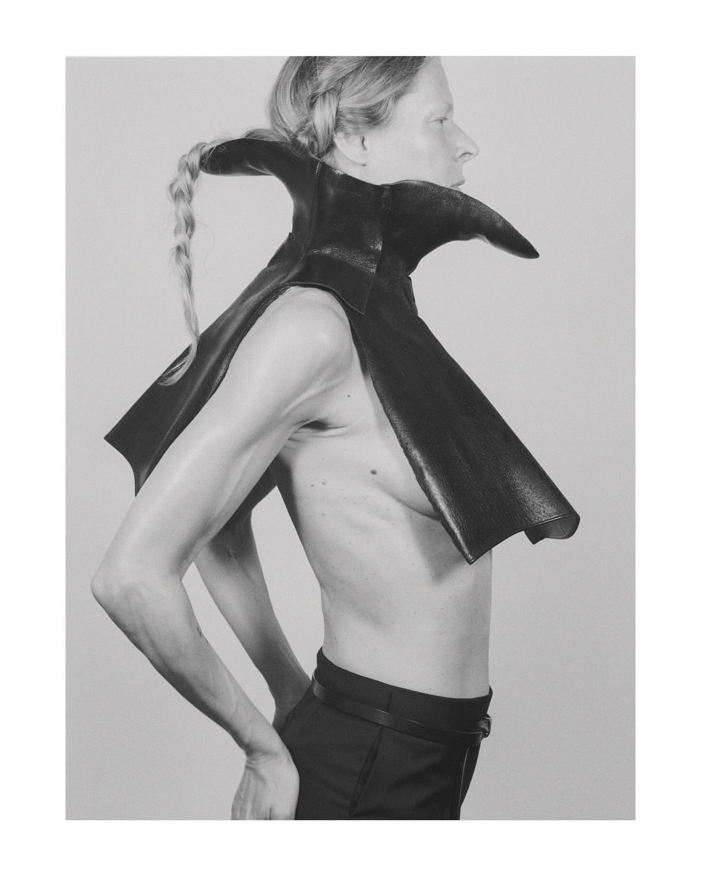 Profile shot of an androgynous person with a blonde braid wearing a black leather cape with a high collar against a grey studio background.