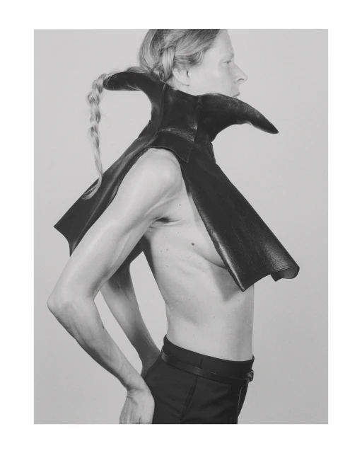 Profile shot of an androgynous person with a blonde braid wearing a black leather cape with a high collar against a grey studio background.