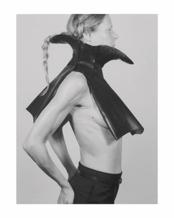 Profile shot of an androgynous person with a blonde braid wearing a black leather cape with a high collar against a grey studio background.