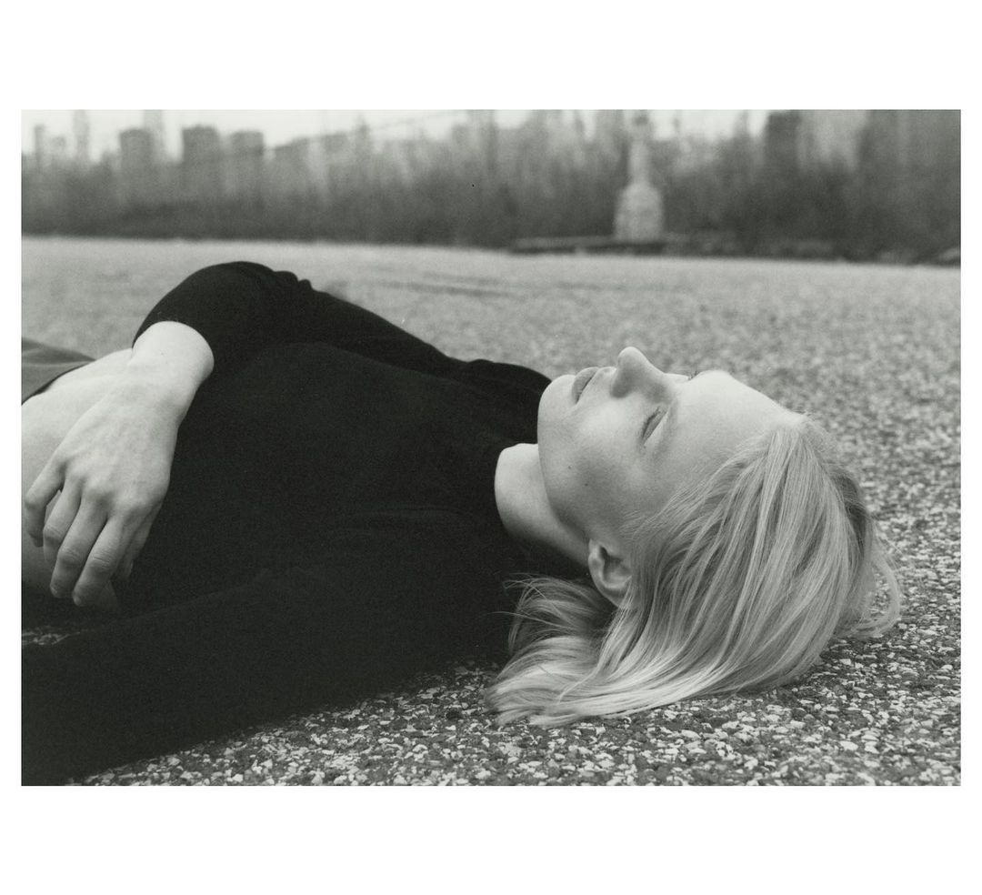 Monochrome portrait of a blonde woman lying down outdoors, with urban buildings and the Statue of Liberty visible in the background.