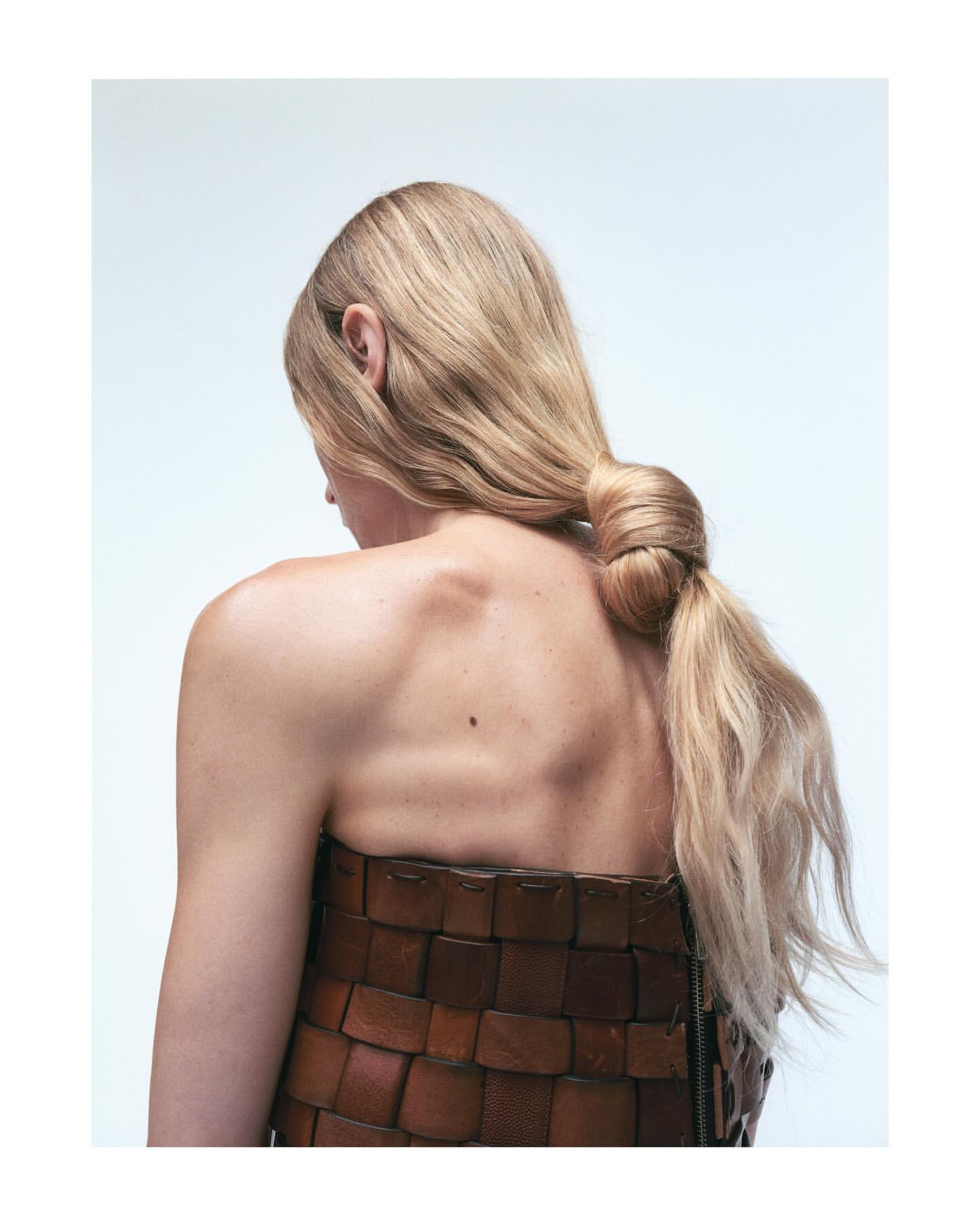 Blonde woman's back view showcases a textured brown woven garment, her hair in a tied ponytail, set against a soft, clean background.