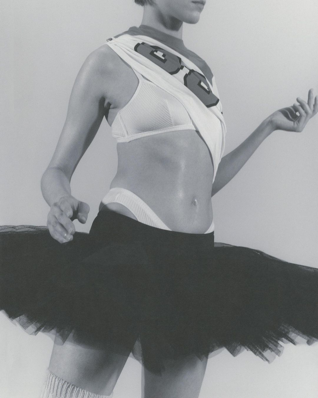 Monochrome fashion shot of a slender woman in a white bra, black tutu, and knee-high socks posing against a neutral background.