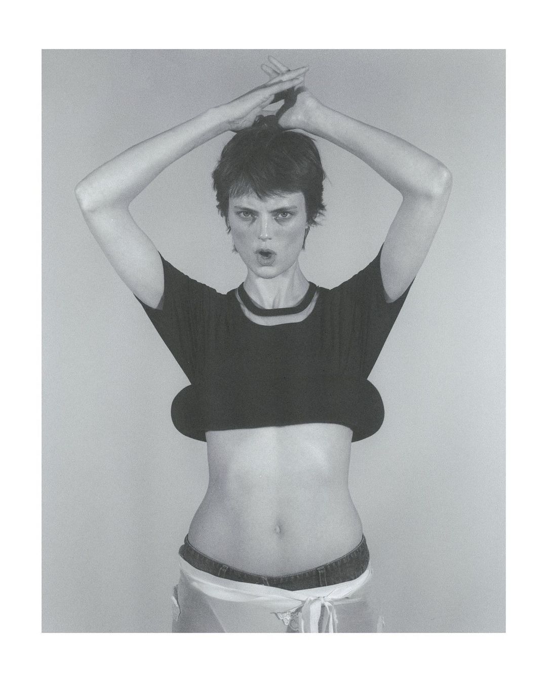 Monochrome portrait of a woman with short hair, arms raised above her head, midriff bared, and mouth open, posing against a gray backdrop.