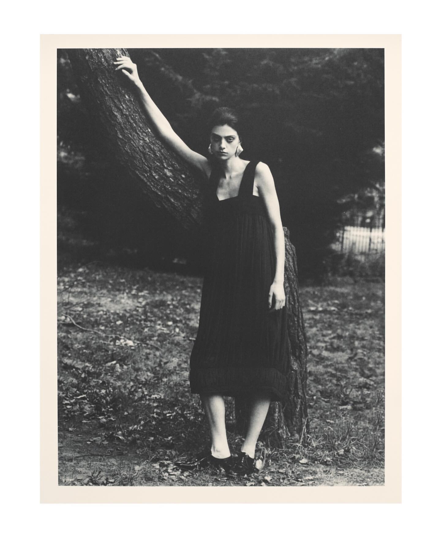 A solitary woman in a black dress poses beside a tree in a moody outdoor fashion shot with monochromatic tones and natural light.