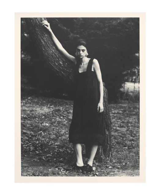 A solitary woman in a black dress poses beside a tree in a moody outdoor fashion shot with monochromatic tones and natural light.