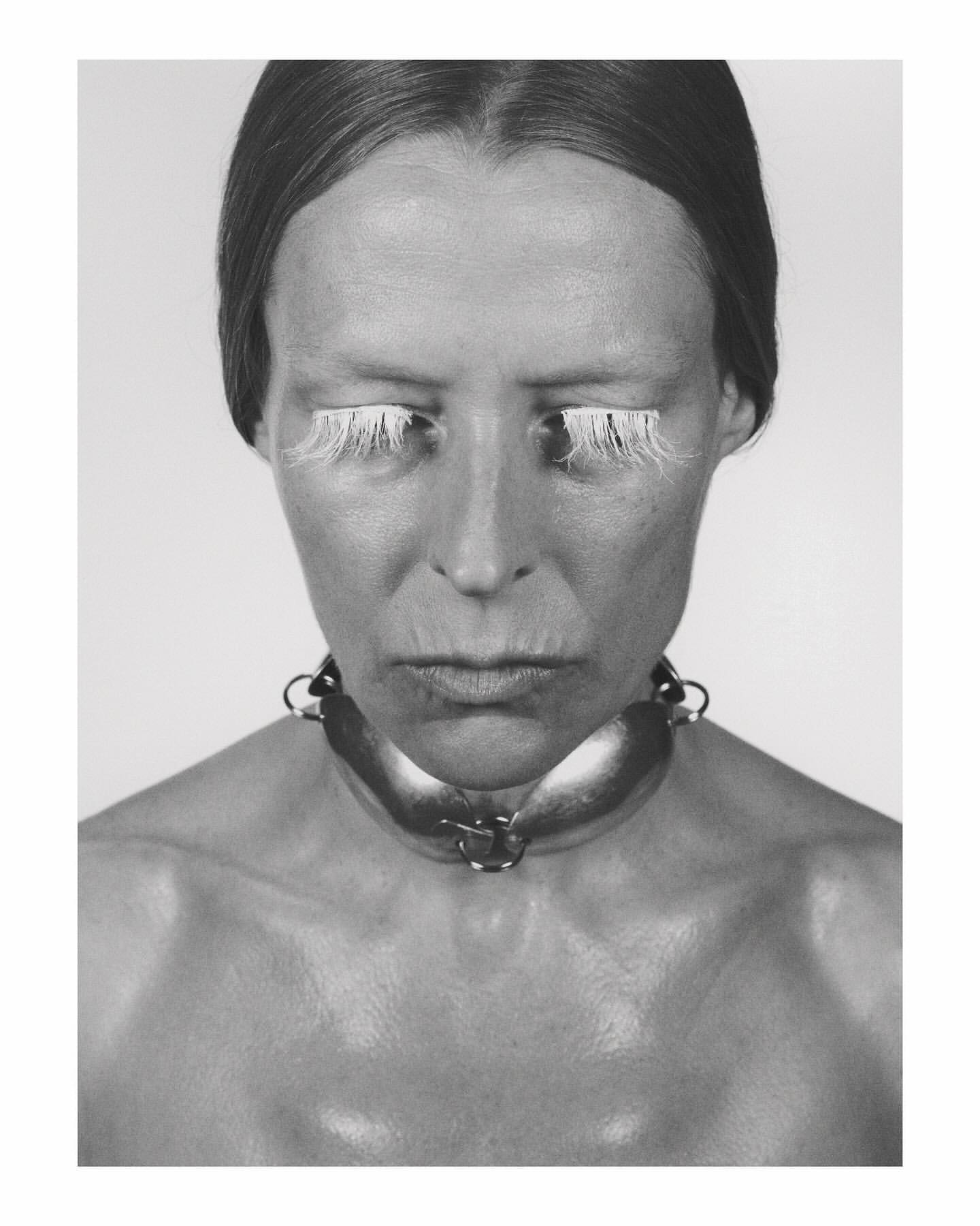 Monochrome portrait of a woman with exaggerated white eyelashes and a wide metal collar, set against a neutral background.