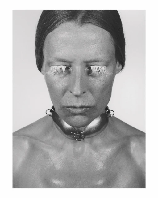 Monochrome portrait of a woman with exaggerated white eyelashes and a wide metal collar, set against a neutral background.