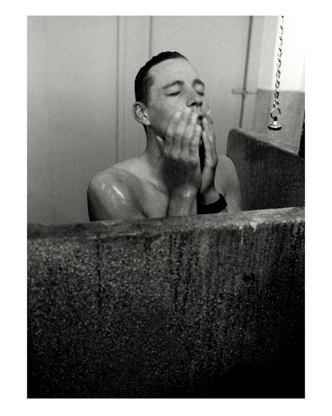 Monochrome portrait of a shirtless man washing his face in a concrete sink, eyes closed, standing in an indoor bathroom setting.