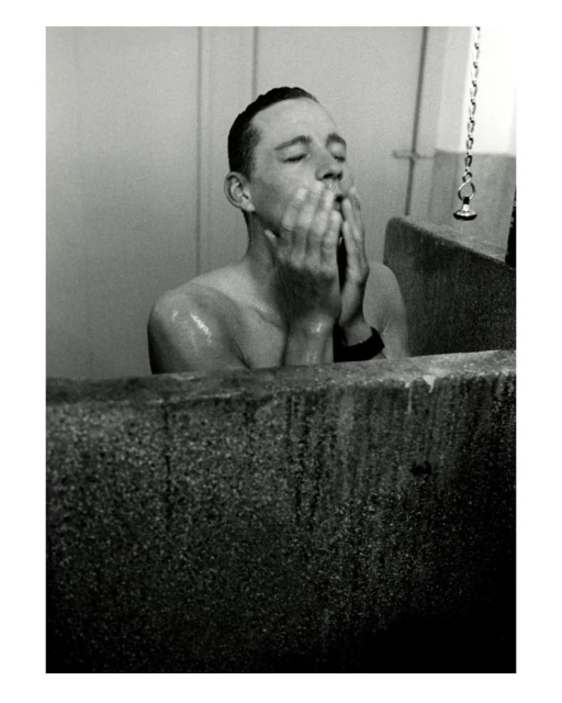 Monochrome portrait of a shirtless man washing his face in a concrete sink, eyes closed, standing in an indoor bathroom setting.