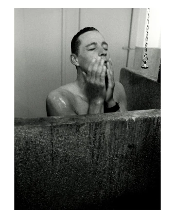 Monochrome portrait of a shirtless man washing his face in a concrete sink, eyes closed, standing in an indoor bathroom setting.