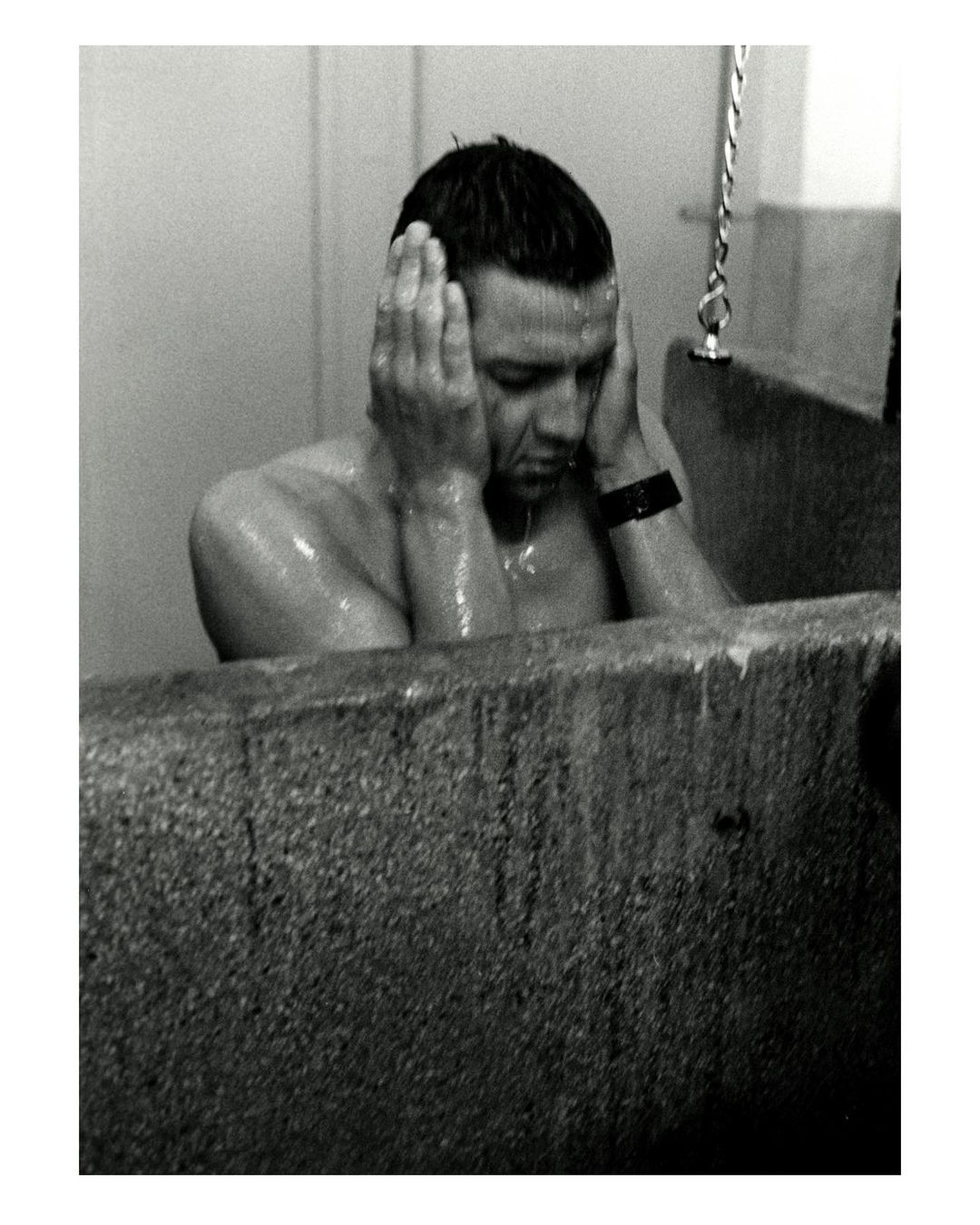 Black and white image of an adult male in a bathtub, with hands covering his ears, facing forward in an indoor bathroom setting.