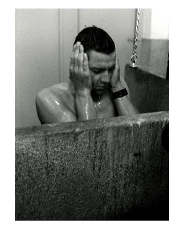 Black and white image of an adult male in a bathtub, with hands covering his ears, facing forward in an indoor bathroom setting.
