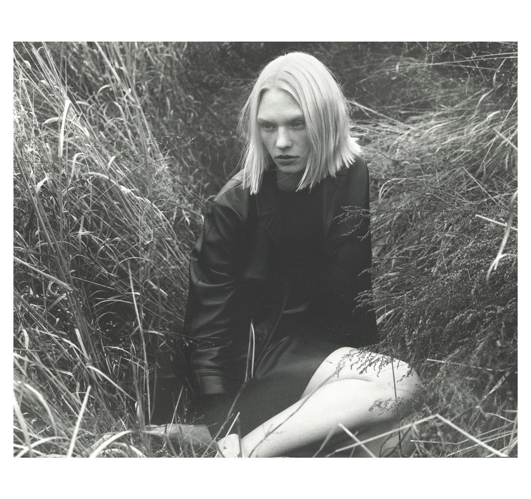 Black and white portrait of a person with short blonde hair and a black jacket sitting in tall grass in an outdoor setting.