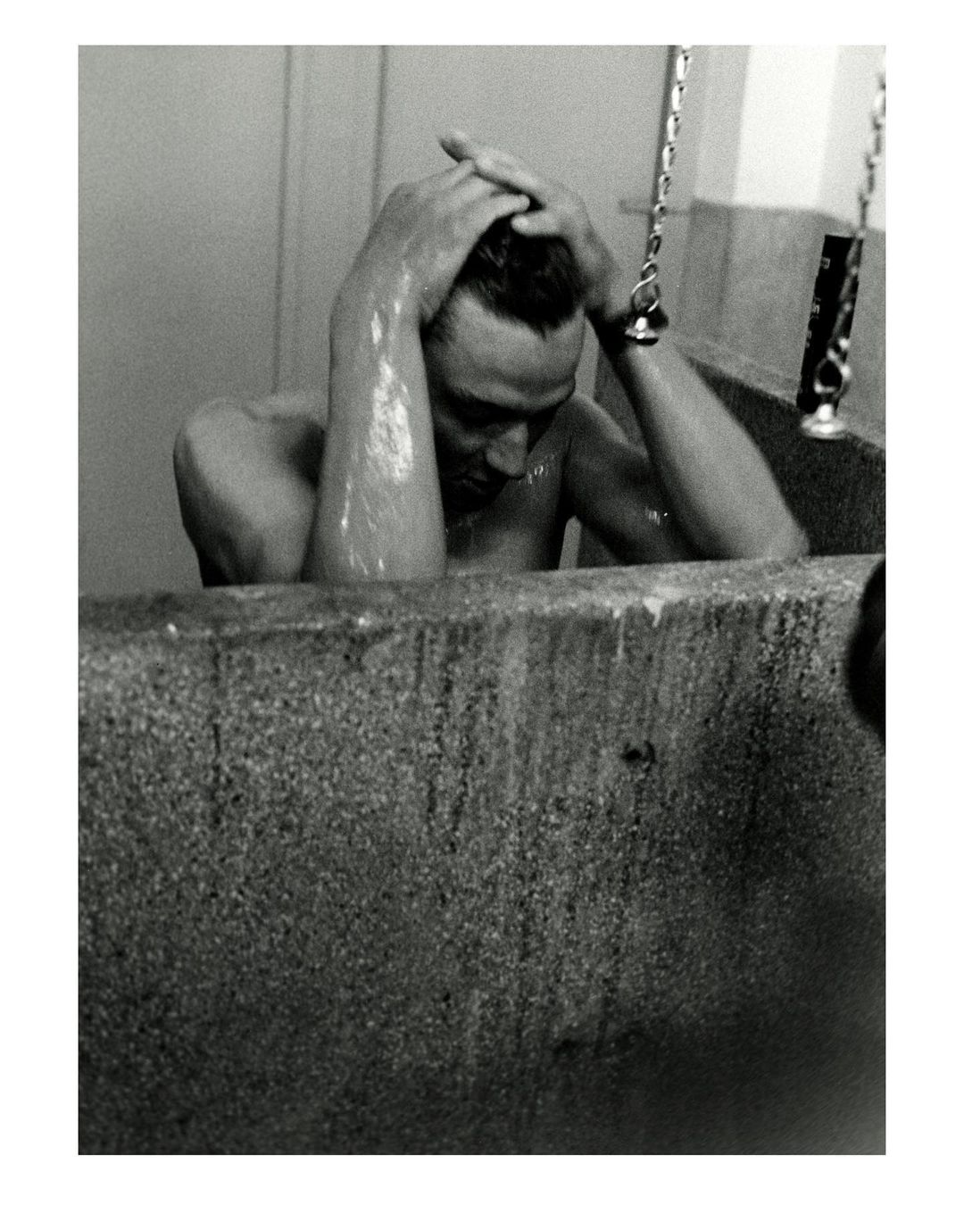 Black and white portrait of an adult male sitting in a bathtub, holding his head, photographed indoors with a close-up camera angle.